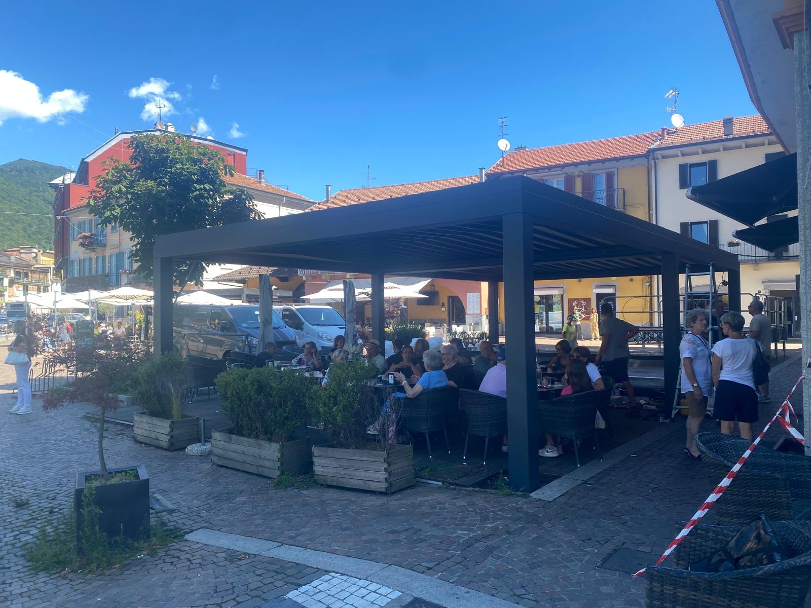 Dehor con struttura scura, lati aperti e tavoli occupati, in una piazza